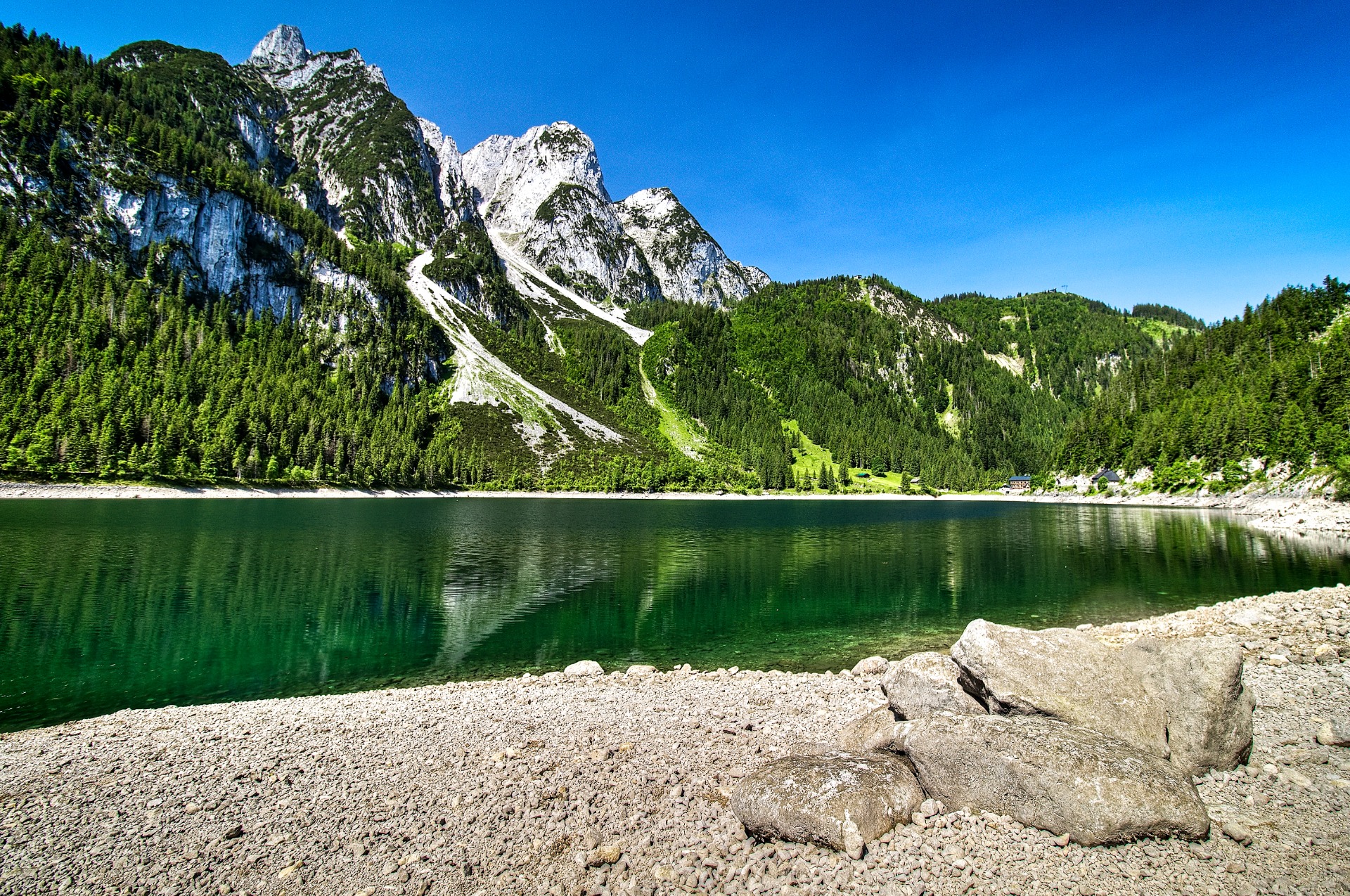 Jezero s klidnou hladinou, skalnaté Alpy v Rakousku a husté lesy pod modrou oblohou.